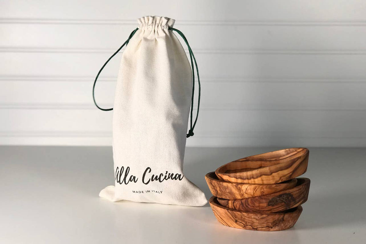 Cotton drawstring gift bag printed Alla Cucina, Made in Italy, beside a stack of the four olivewood condiment bowls