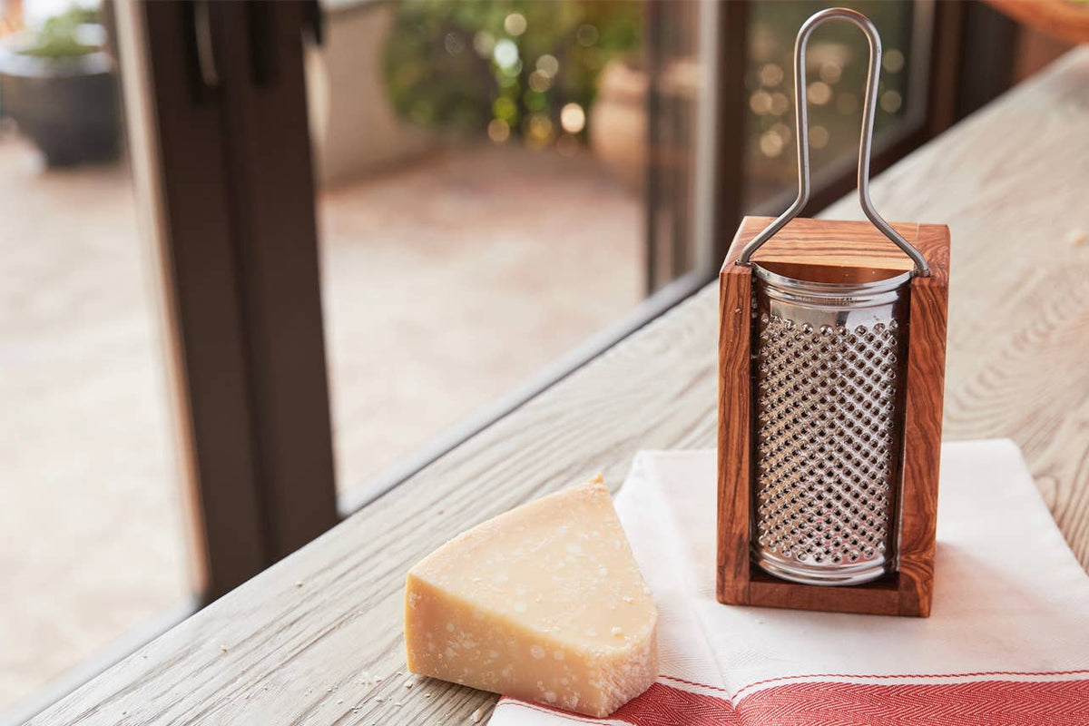 Olivewood box cheese grater standing upright on a linen napkin beside a wedge of Parmigiano-Reggiano