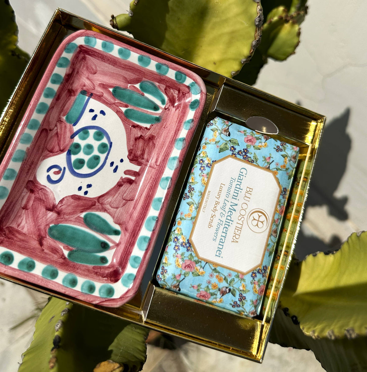 Giardini Mediterranei gift box open in sunlight: a Tomato Leaf and Flowers body scrub beside a hand-painted Vietri ceramic soap dish with a pink and green fish design
