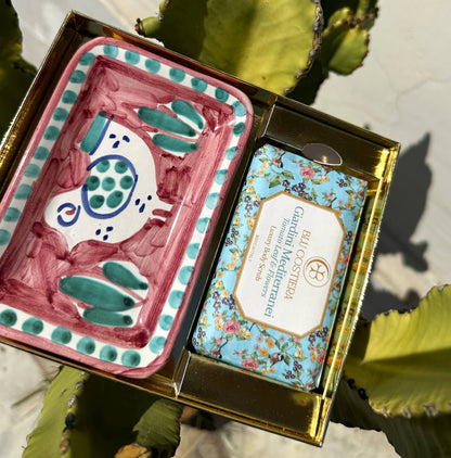 Giardini Mediterranei gift box open in sunlight: a Tomato Leaf and Flowers body scrub beside a hand-painted Vietri ceramic soap dish with a pink and green fish design