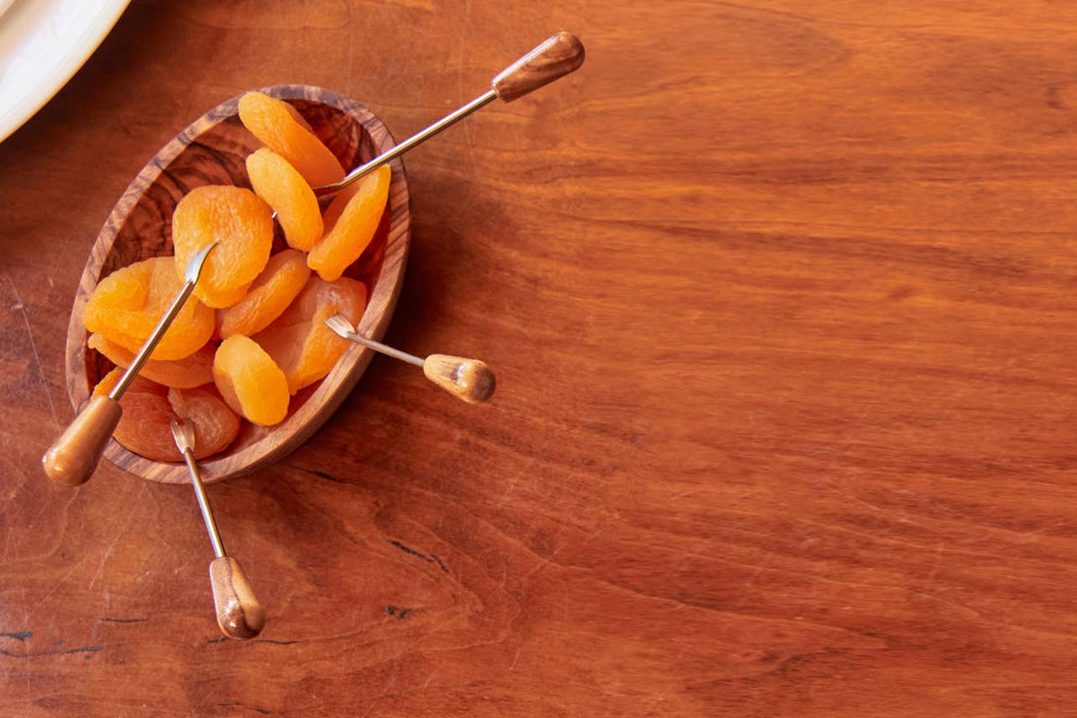 Overhead view of a single olivewood condiment bowl holding dried apricots with olivewood-handled cocktail picks