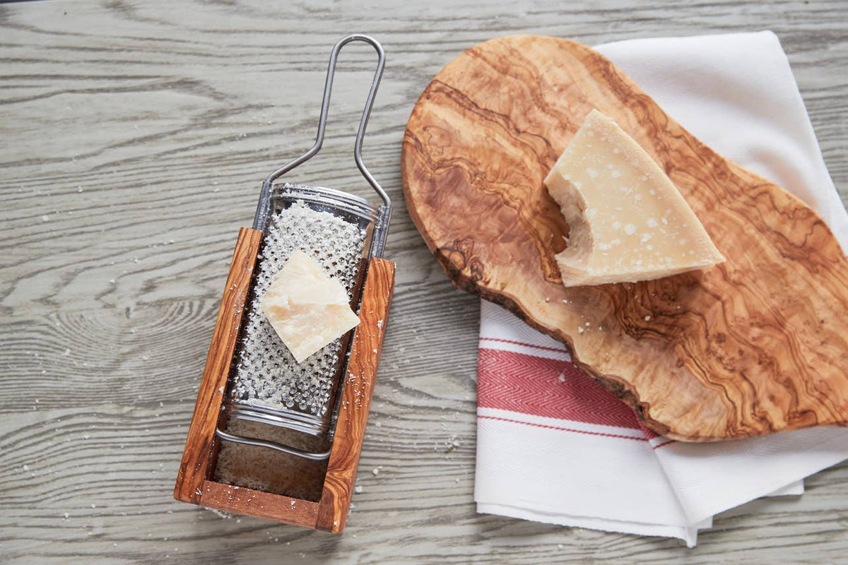 Overhead view of the olivewood box cheese grater beside an olivewood serving board, Parmigiano wedge and red-striped kitchen towel