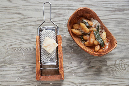 Olivewood box cheese grater beside an olivewood bowl of gnocchi, with a wedge of Parmigiano resting on the grater plate