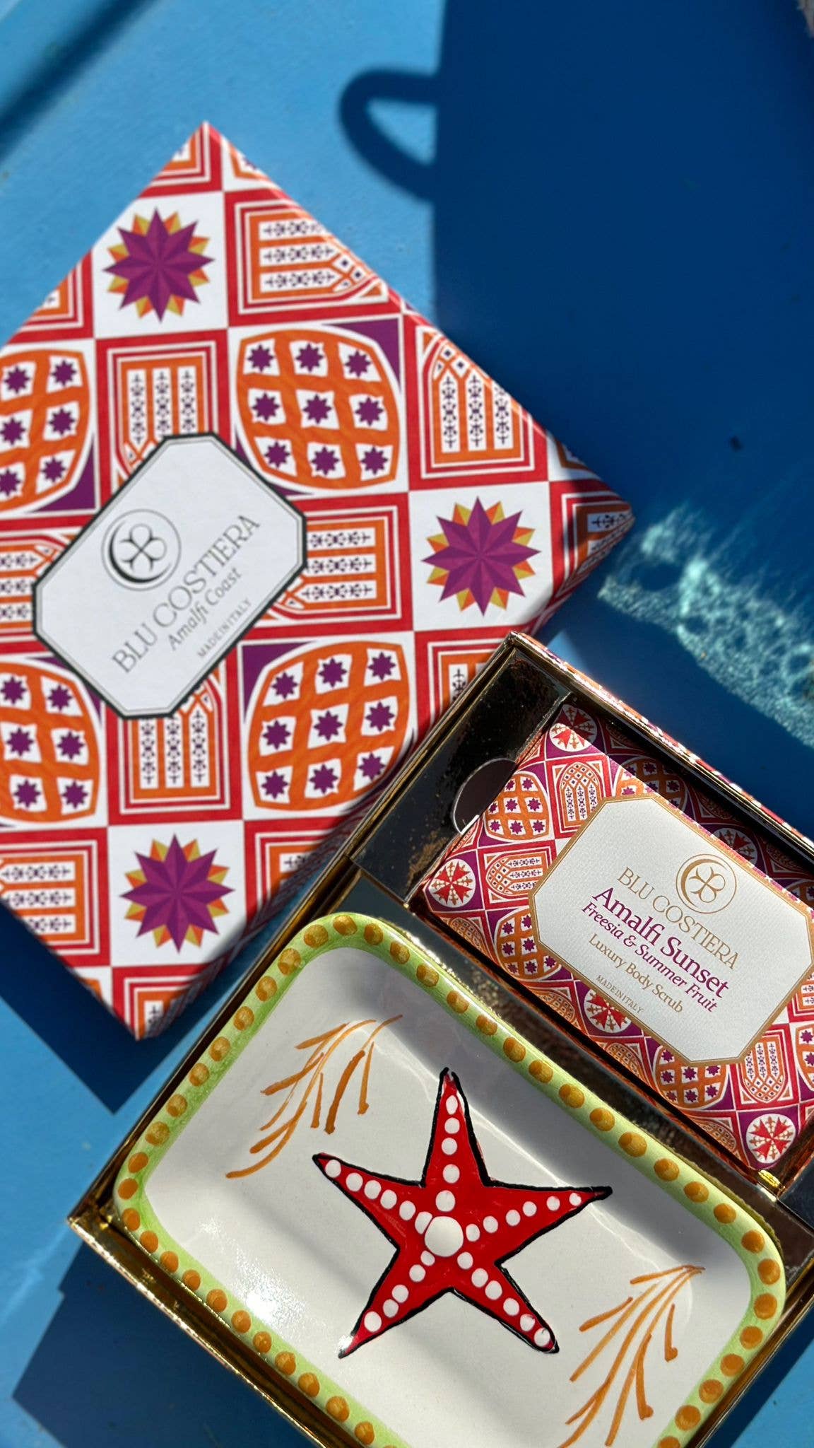 Blu Costiera Amalfi Sunset gift box photographed on blue painted wood, lid printed with orange, red and magenta maiolica tilework, open base showing the body scrub beside a hand-painted Vietri ceramic dish with a red starfish