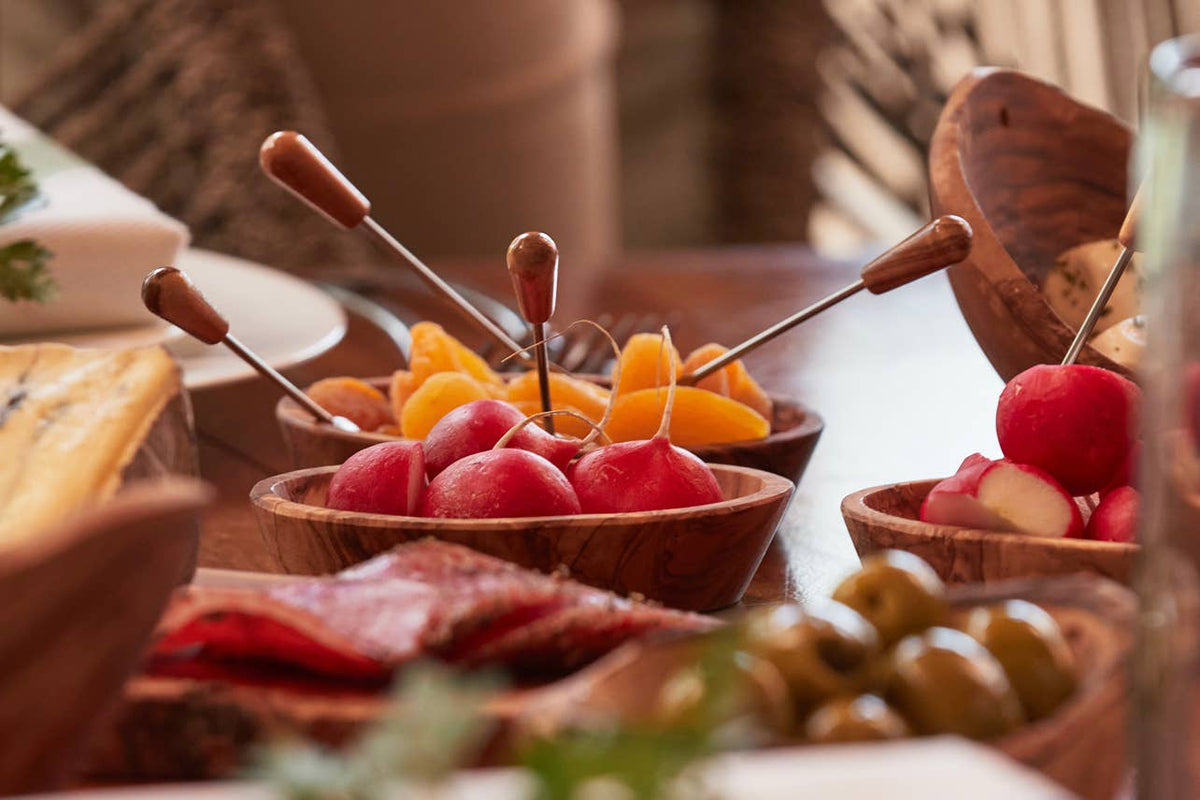 Olivewood condiment bowls holding radishes and dried apricots on an aperitivo table with cured meats and olives