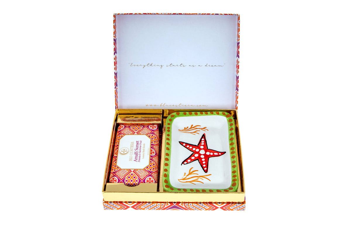 Amalfi Sunset gift box open, lid printed 'Everything starts as a dream', holding the Freesia and Summer Fruit body scrub beside a hand-painted Vietri ceramic soap dish with a red starfish, orange coral and a green dotted rim