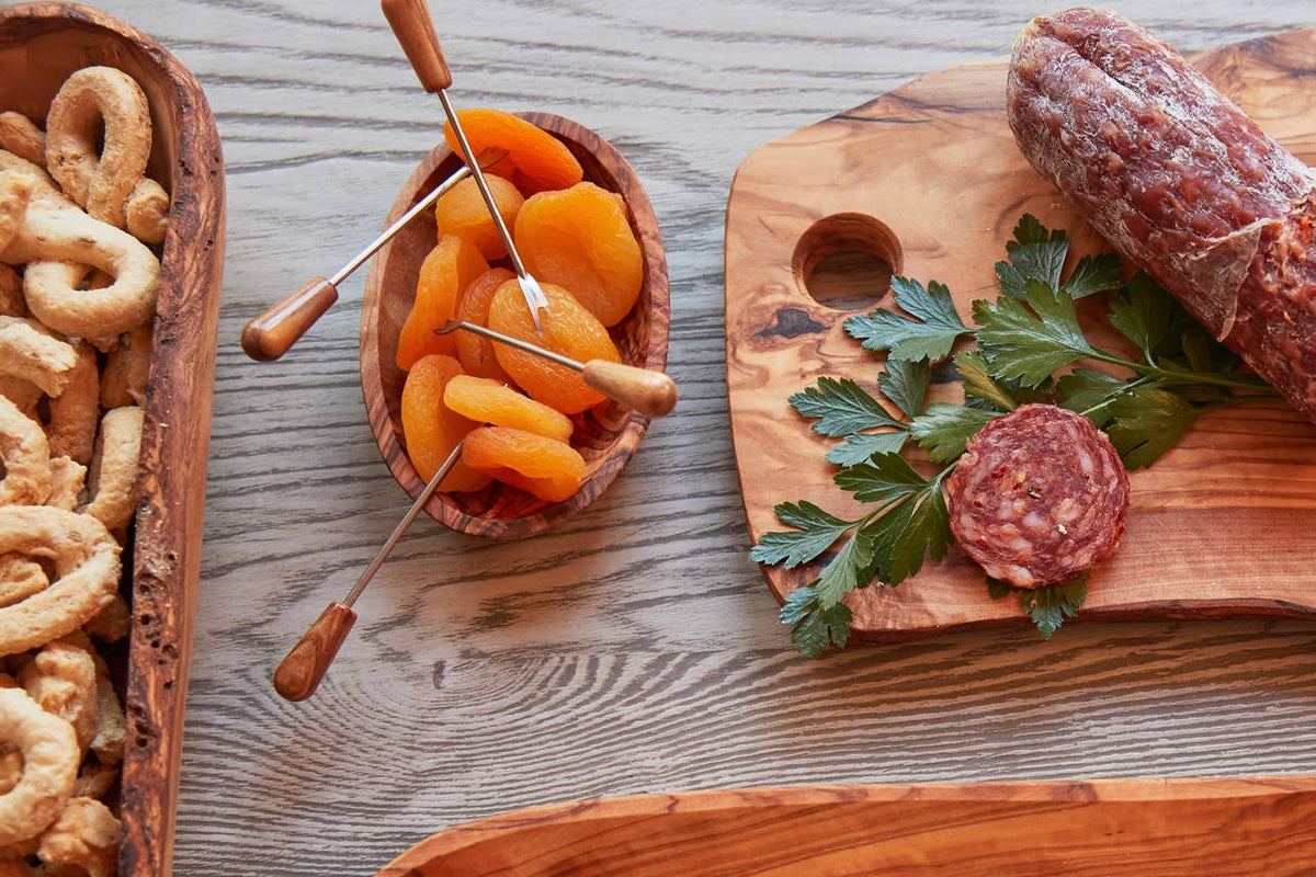 Olivewood condiment bowl of dried apricots beside taralli, salami and parsley on an olivewood serving board