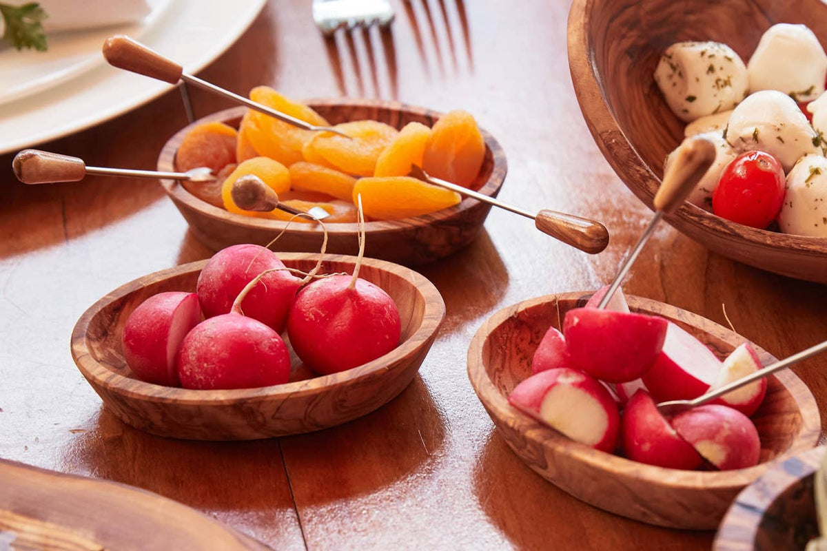 Several olivewood condiment bowls filled with radishes, apricots and mozzarella on a wooden aperitivo table with cocktail picks