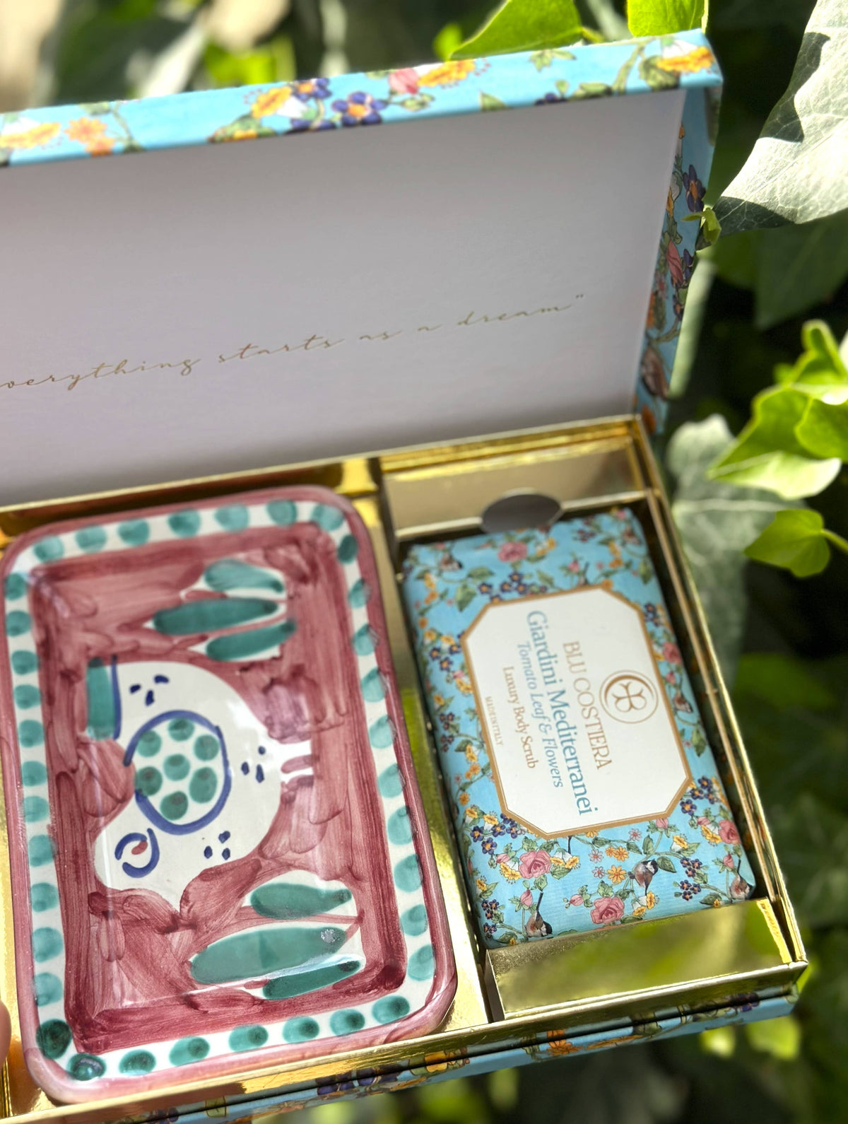 Open Giardini Mediterranei gift box held against garden foliage, showing the Blu Costiera body scrub and a hand-painted Vietri ceramic soap dish with a pink and teal fish