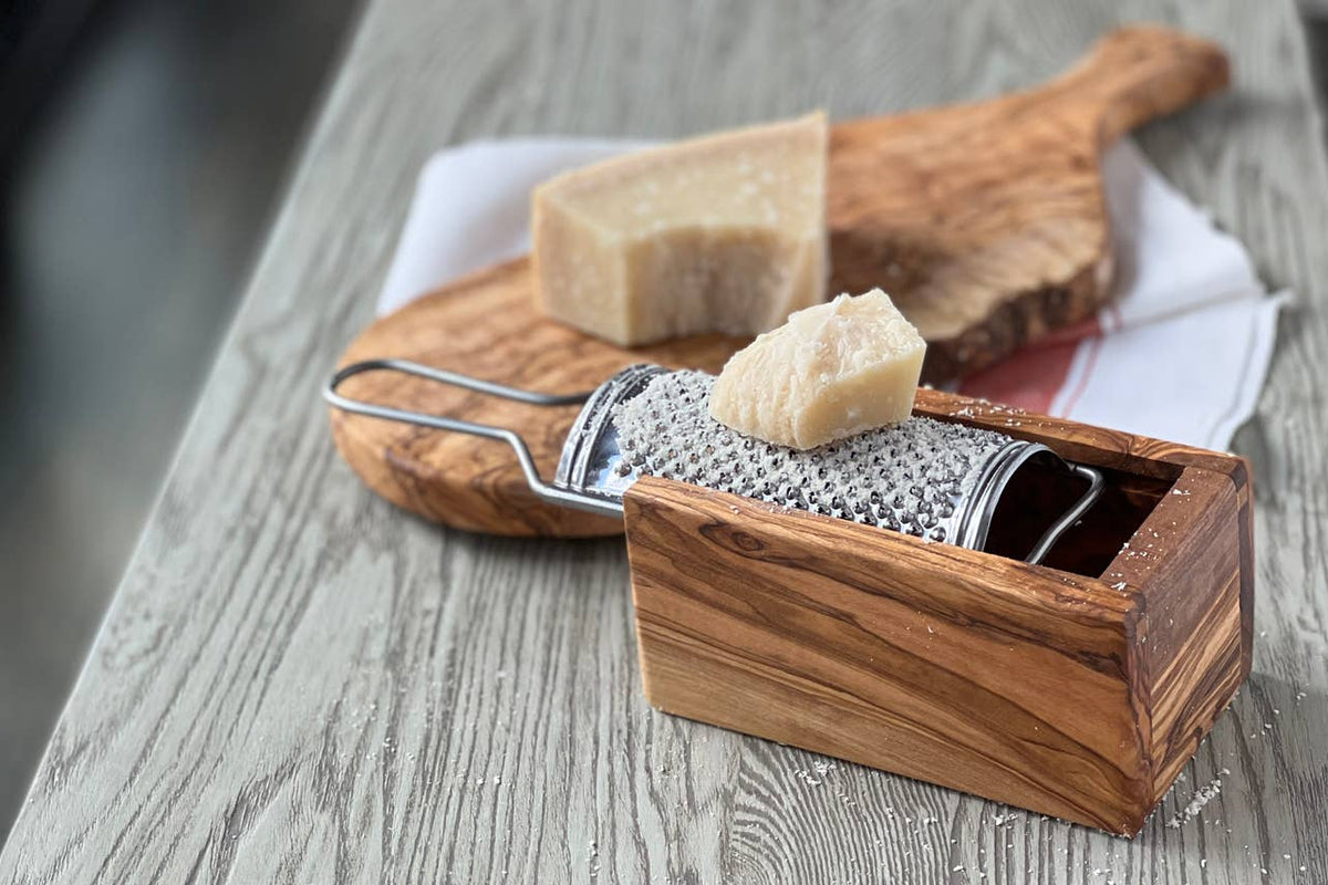 Olivewood cheese grater with grated Parmigiano in the box, styled with an olivewood board and wedges of hard cheese